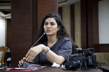 A person with long hair speaks into a microphone at a table, with a video camera and papers beside them.