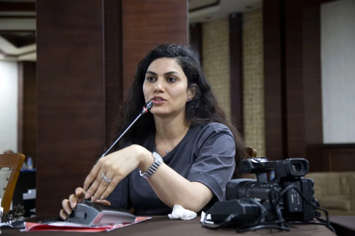 A person with long hair speaks into a microphone at a table, with a video camera and papers beside them.