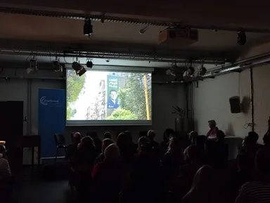 An audience seated in a darkened room watches a projected film showing a street mural of a man's face beside an Arabic slogan, with a blue elbarlament banner standing to the left of the screen.