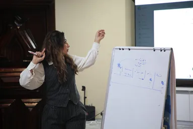 A woman points at a whiteboard with Arabic handwriting while a presentation is displayed on a screen behind her.