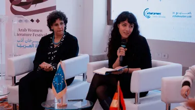Two women seated during a panel discussion, one speaking into a microphone while holding an open notebook, beside a banner reading "Arab Women Literature Festival" and a screen showing the elbarlament logo.
