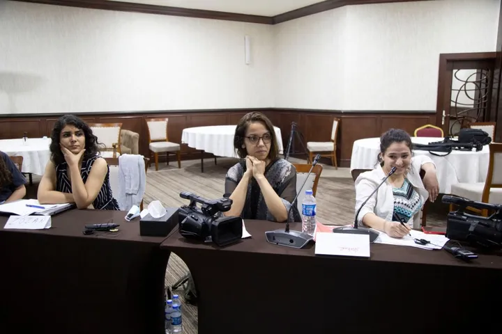 Three women sit behind a conference table with video cameras, microphones and water bottles, in a hotel meeting room with round tables in the background.