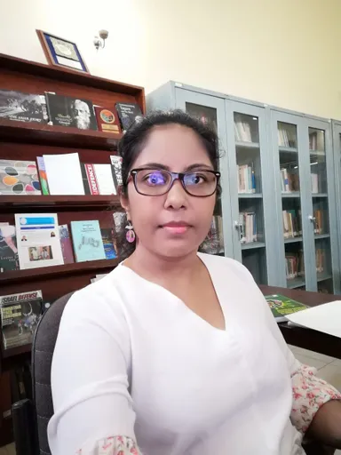 A woman wearing glasses sits in an office in front of wooden bookshelves and a glass-fronted cabinet filled with books.