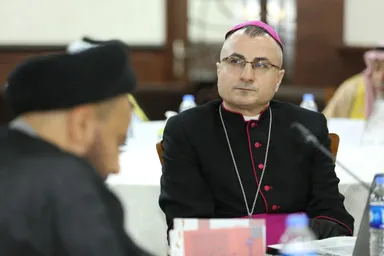 A man wearing glasses, a pink skullcap and a white collar with red trim sits at a conference table, with another man blurred in the foreground.