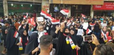 A crowd of protesters, including women in black robes with red-white-black sashes, wave national flags on a street lined with shops.