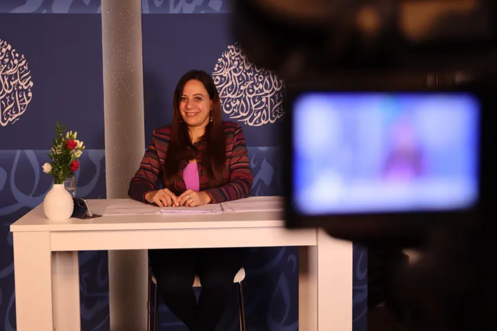 A woman sits smiling at a white table in a TV studio decorated with circular Arabic calligraphy panels, with a camera and screen visible in the foreground.
