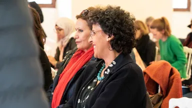 Two women sitting close together, smiling as they listen during an event, with other attendees seated behind them.