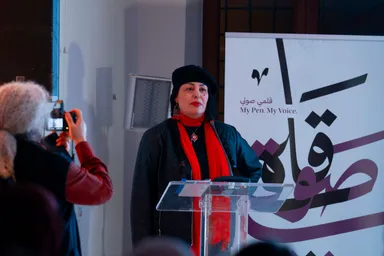 A woman in a black hat and red scarf speaks at a clear lectern in front of a banner reading "My Pen. My Voice." and Arabic text, while someone photographs her from the crowd.