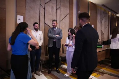 A group of people stand talking, with a sign reading "ذي قار" on the wall, chairs and a table with cups in the background.
