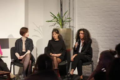 Three women sit on a panel stage in front of a potted plant and a white brick wall, one speaking into a microphone, an audience seated in the foreground.
