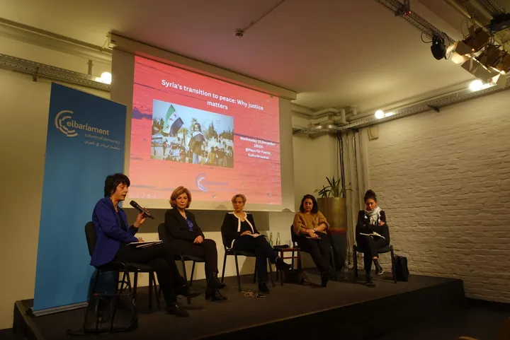 Five women sit in a panel discussion on a stage, with a screen behind them reading "Syria's transition to peace: Why justice matters".