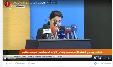 Facebook Live video screenshot of a woman speaking at a podium with several branded microphones, and a Kurdish caption bar reading "Recorded live".