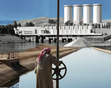 A man in a long robe and red-and-white headscarf turns a valve wheel on a canal in front of a concrete dam building with four tall cylindrical towers.