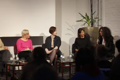 Five people sit on a panel stage in front of a potted plant and a white brick wall, one woman speaking into a microphone, water bottles on tables in front, an audience seated in the foreground.