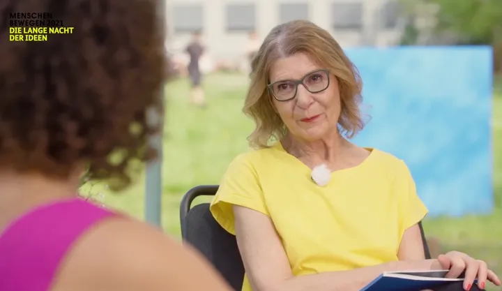 A woman wearing glasses and a yellow top is interviewed outdoors, with the on-screen text "MENSCHEN BEWEGEN 2021 DIE LANGE NACHT DER IDEEN" in the corner.
