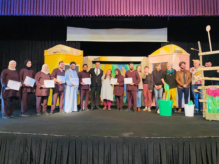 Group photo on a stage of performers in costume holding certificates, alongside other people, in front of theater set pieces and a curtain backdrop.