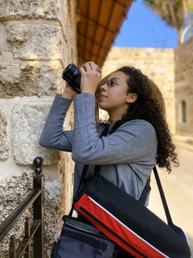A woman with curly hair holds a camera to her eye in a stone archway, a camera bag slung over her shoulder.