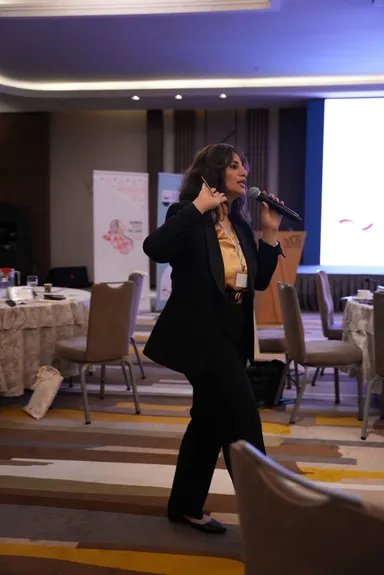 A woman walks while speaking into a microphone and holding a pen, with banners and a blank screen behind her and round tables in the foreground.