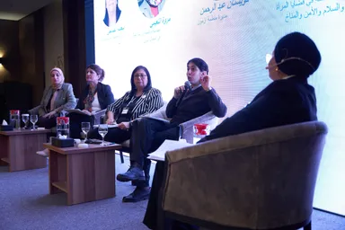 Five people sit in armchairs on a stage, one speaking into a microphone with a hand raised, in front of a screen displaying Arabic text with speaker names and session details.