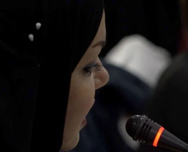 Close-up profile of a person in a headscarf next to a microphone with a lit indicator ring, in a dim room.