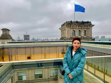 A woman in a teal coat stands on a rooftop terrace with a domed tower and an EU flag in the background.
