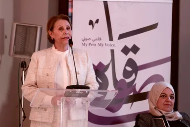 A woman speaks into a microphone at a lectern, in front of a banner reading "My Pen. My Voice.", with another woman seated nearby.