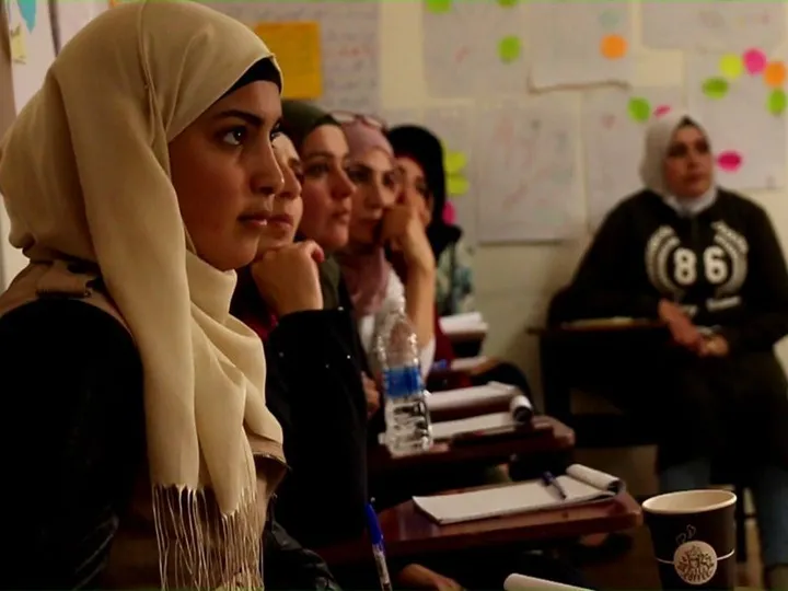 Row of young women seated at wooden school desks with notebooks, pens and a water bottle, listening in a classroom whose wall is covered with handwritten flipchart sheets.