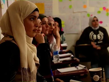 Row of young women seated at wooden school desks with notebooks, pens and a water bottle, listening in a classroom whose wall is covered with handwritten flipchart sheets.