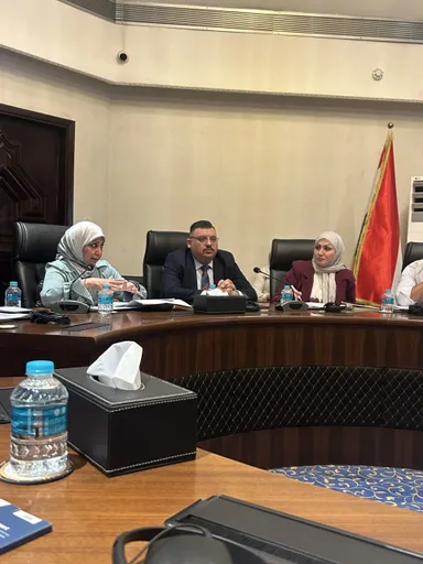 A woman speaks into a microphone at a curved conference table with two other people, water bottles and papers in front of them, a red flag standing behind.