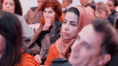 A woman wearing an orange headscarf sits among a seated audience, looking to the side.