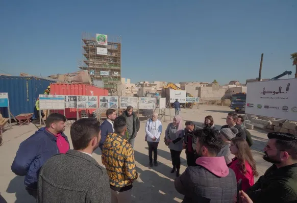 Group of people standing outdoors at a reconstruction site with information boards and scaffolding, near a banner reading "Reviving The Spirit Of Mosul".