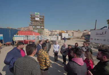 Group of people standing outdoors at a reconstruction site with information boards and scaffolding, near a banner reading "Reviving The Spirit Of Mosul".