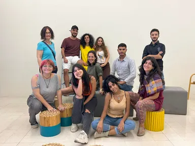 A group of people posing together indoors, some standing and some seated or kneeling on wooden stools, against a plain wall.
