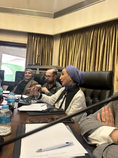 A woman in a blue headscarf gestures with a pen while speaking, seated at a table with three other people, papers and water bottles in front of them, gold curtains behind.