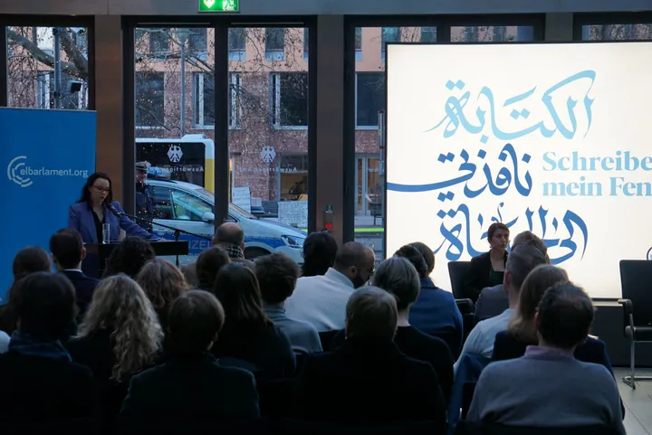 An audience listening to a woman speaking at a lectern beside an "elbarlament.org" banner, with a large screen showing Arabic calligraphy and German text in the background.