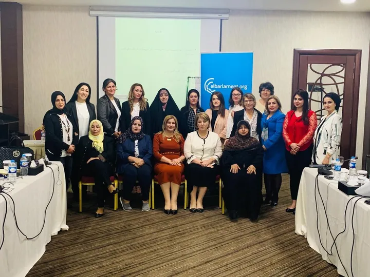 A group of women standing and seated for a photo in a conference room, with a blue banner reading "elbarlament.org" and a screen showing text in the background.