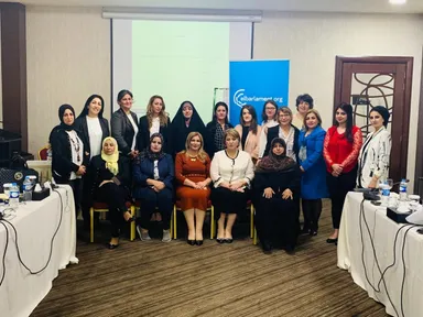 A group of women standing and seated for a photo in a conference room, with a blue banner reading "elbarlament.org" and a screen showing text in the background.