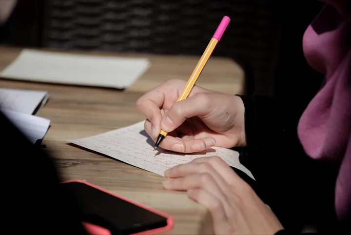 A hand writing on paper with a pencil at a wooden table, a phone and notebooks nearby.