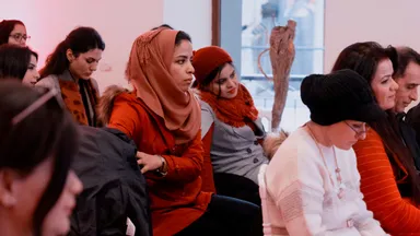 A group of people sitting in rows indoors, wearing colorful scarves and hats, with a window visible behind them.