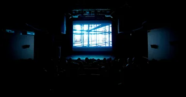 Audience seated in a dark auditorium watching a bright screen showing scaffolding against the sky.