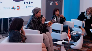 Four women sit together in a panel discussion, one holding a microphone and a book, with a Tunisian flag on the table and a banner reading "WORLD ART CONNECTION" behind them.