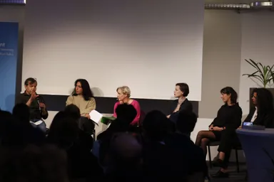 Six people sit on a panel stage in front of a blank projection screen and a plant, a banner partly showing "elbarlament cultures of democracy" text, an audience seated in the foreground.