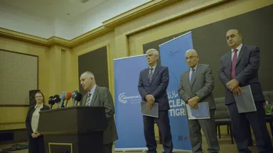 A man speaks at a podium with several branded microphones, a woman standing beside him and three men behind, in front of "elbarlament" and "Clean Tigris" banners.