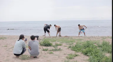 Three men digging in sand near a shoreline while two people crouch nearby filming with a phone and a camera on a tripod.