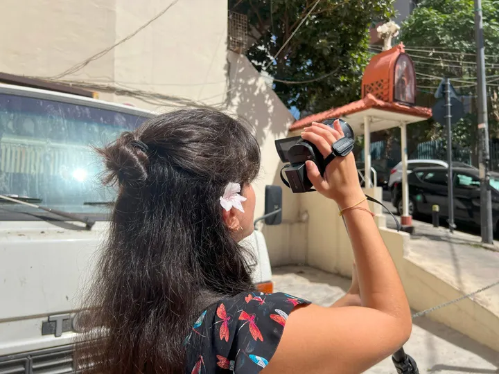 A person holds up a video camera to film a street scene with a parked van and a small wall-mounted shrine.
