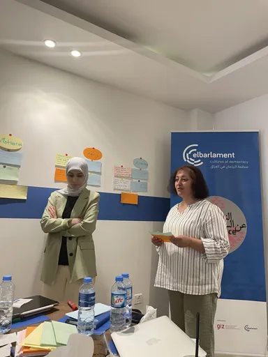 A woman with arms crossed stands beside sticky notes labelled "Motivation" and "Skills" while another woman speaks near a banner reading "elbarlament cultures of democracy" and "Women Talking Peace".