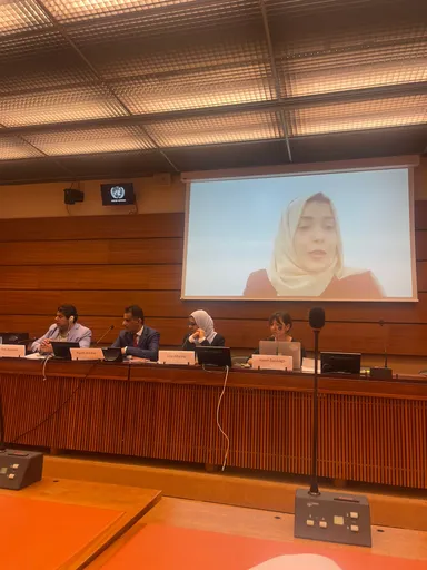A person's face is shown on a large screen during a panel discussion, with several people seated at a table with laptops and nameplates in a conference room.