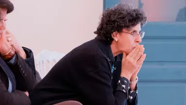 A woman sitting with clasped hands in front of her mouth, listening attentively.