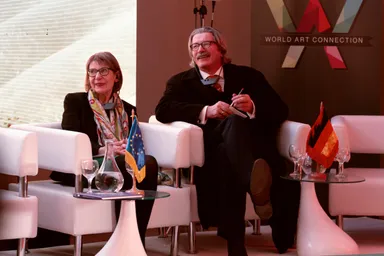 Two people sit in white armchairs on a stage, with European Union and German flags on side tables and a banner reading "WORLD ART CONNECTION" behind them.