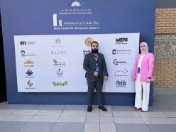 A man and a woman stand in front of a large board displaying numerous organisation logos in Arabic and English.